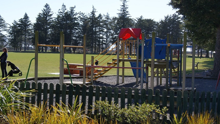 A picture of the Kirwee Domain playground with two women in the distance and a sports field behind