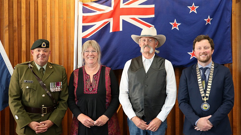 A man in Army dress uniform, a blonde woman in a black top, a man in a large white stetson hat with a bushy grey moustache and Mayor Sam Broughton in his mayoral chains stand in front of a New Zealand flag