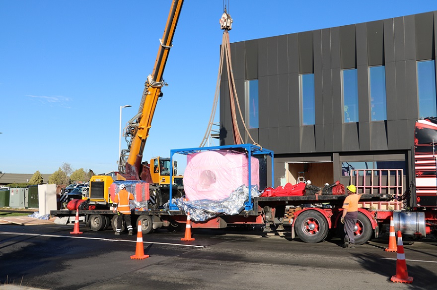 A crane lifts a CRT machine off the back of a flatbed trailer in front of the new Selwyn Health Hub