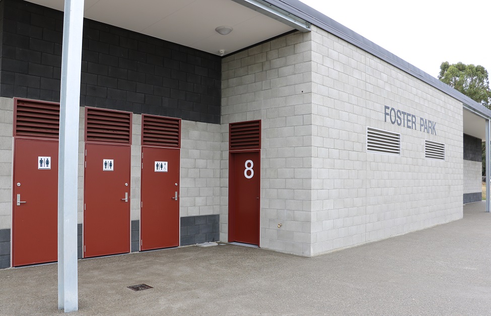 The outside of a breeze block building with three red toilet doors, a red door with a white 8 on it and the sign Foster Park on the wall