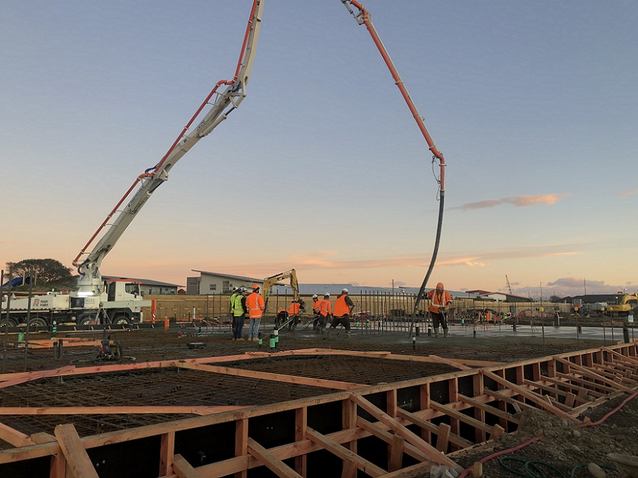 View across work site in early morning, truck with high arching hose pours concrete, spread by workers in hi-vis