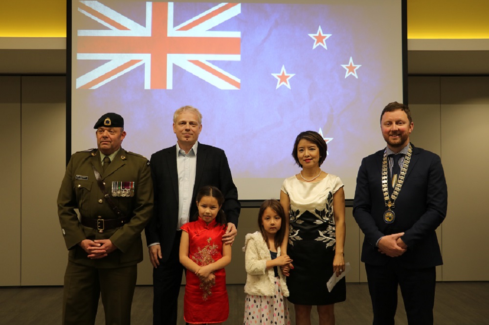 A New Zealand Army officer in full uniform stands next to a tall man with white blonde hair with his arm on the shoulder of a young girl in a red dress, a young girl in a white dress holds the arm of a Chinese woman in a black and white dress standing next to Mayor Sam Broughton wearing his Mayoral chains