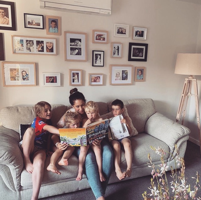 A mum and four young children sit on a couch reading a story and pointing at the pictures