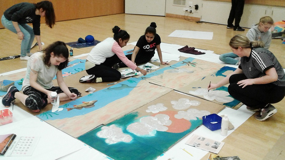 A group of students sitting on the floor painting a nature scene on large wooden boards