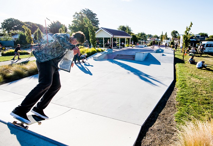 A skateboarder at the top of a shallow half-pipe in a grassed area with people in the paths and grassed area in the background