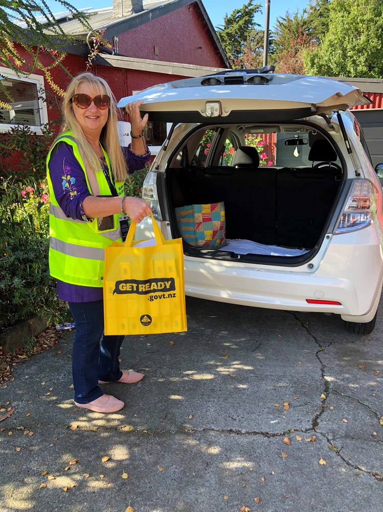 A blonde woman in a yellow high visibility jacket standing at the boot of a car holding a yellow reusable bag with emergency management branding
