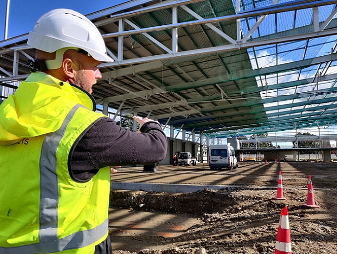 A man in hard hat and high viz jacket points to a roof covering half the roof beams on a construction site