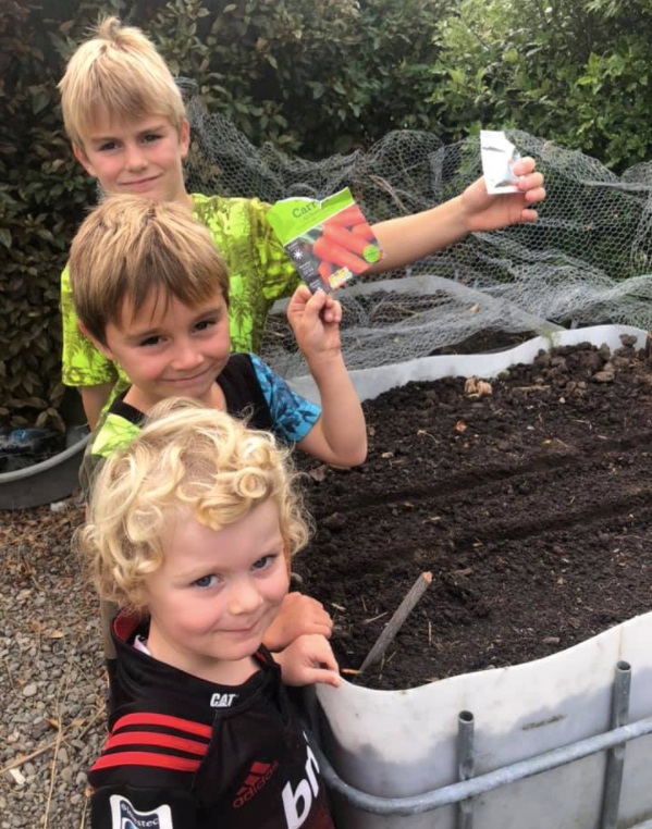 Three blonde children stand in a line holding out seed packets next to a large plastic container filled with earth