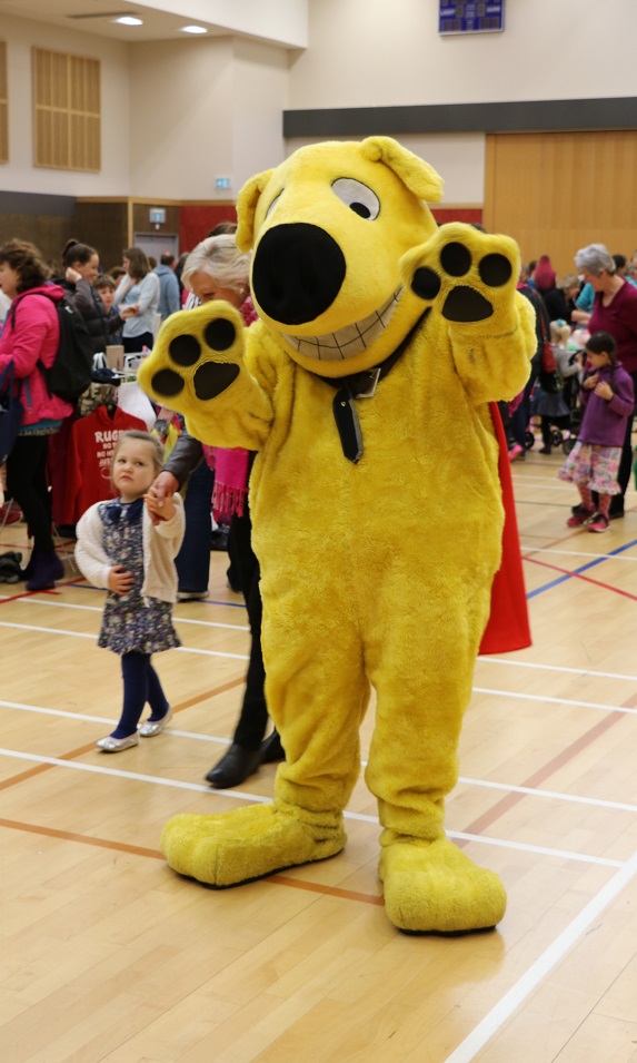 Yellow mascot dog CoolStuff waves at camera in front of children playing in a gym