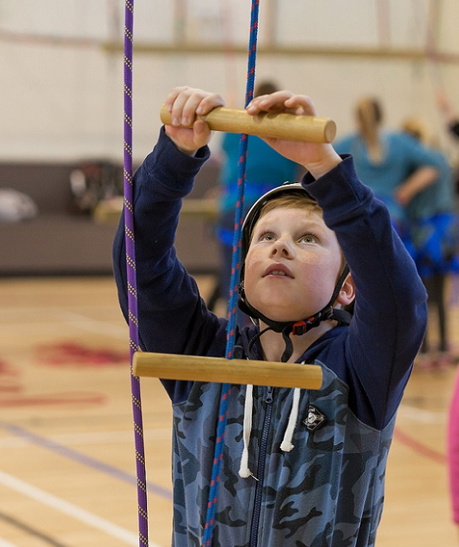 Boy in blue camouflage jersey and white climbing helmet gets ready to climb a rope ladder