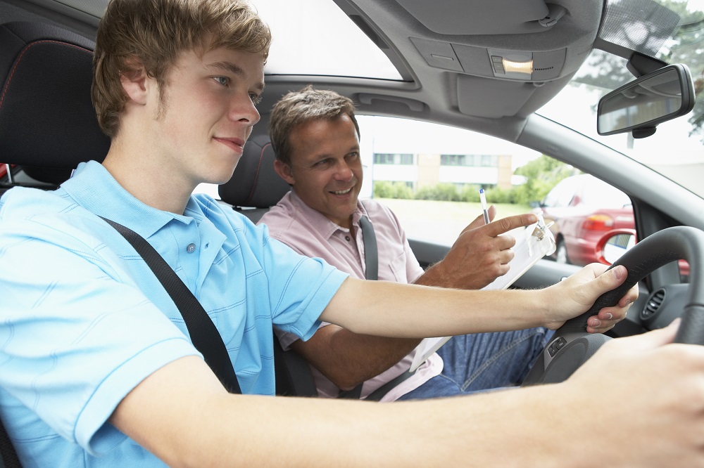 Teenager in a blue polo shirt drives a car while a man with a clipboard and a pen points and gives an instruction