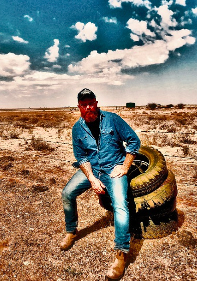 Promotional image of Adam McGrath sitting on a stack of old tires next to a barbed wire fence 