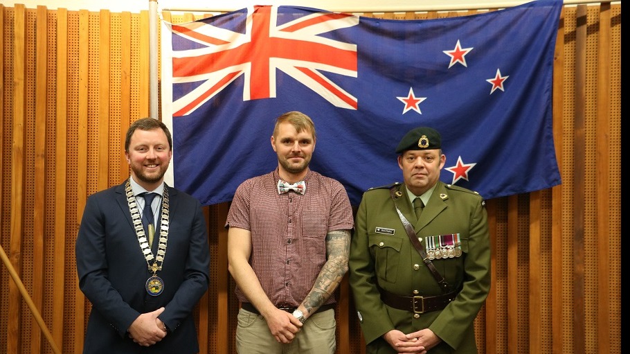 Picture of Mayor Sam Broughton, blonde man in red check shirt and bow-tie and New Zealand Army Major in uniform in front of a New Zealand flag