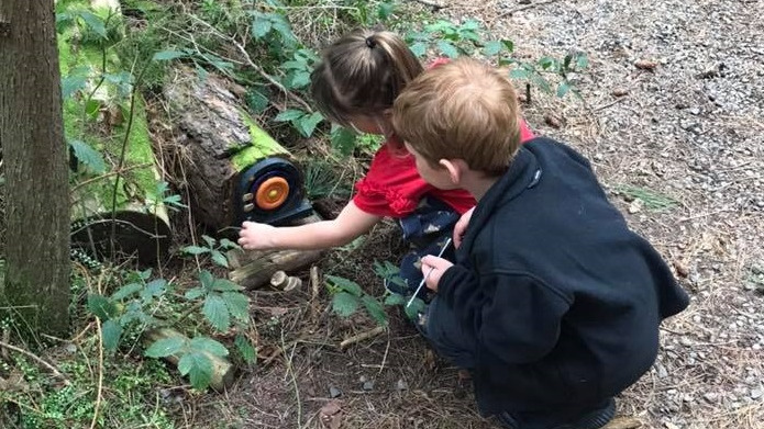 Two children, a boy in a black jumper and a girl in a red shirt crouch down looking at a small black door with round handle attached to a log