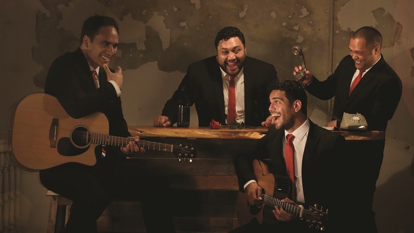 The four members of the Modern Maori Quartet laughing, two members playing guitars in front of a piano and two members behind laughing