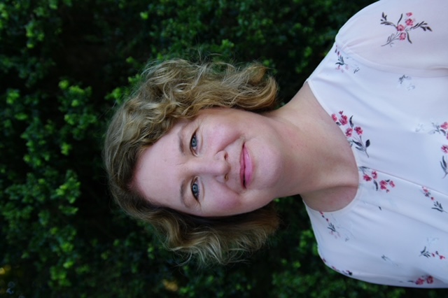 head and shoulder shot of woman in white top with flower pattern, blonde curly hair, standing in front of a green hedge