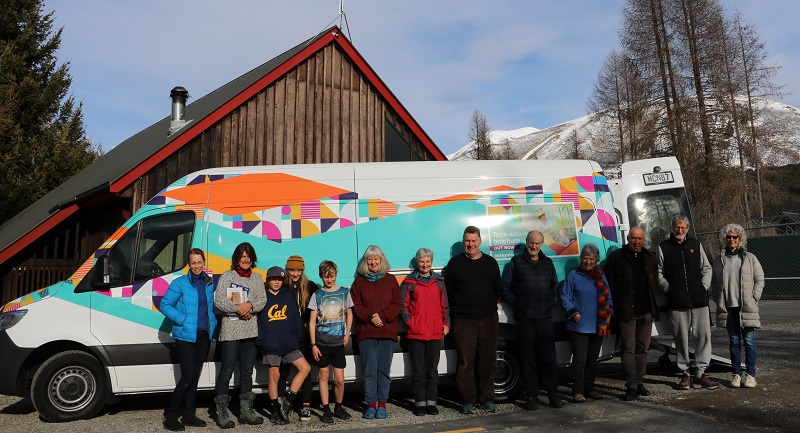 A line of 13 people stand in front of a white van with a blue and orange logo