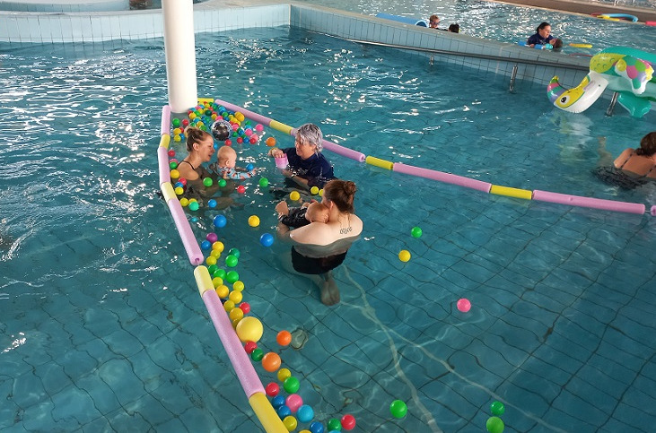 Two mothers with babies and an instructor playing with coloured balls in an area separated by floating foam barriers