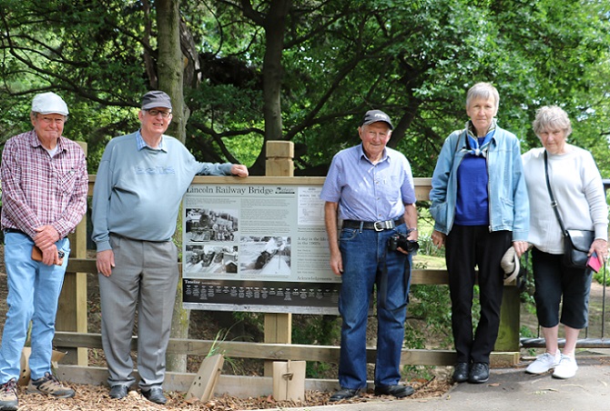 A line up of older people on a bridge either side of a pale green sign saying Lincoln Historic Railway Bridge and showing pictures and information about the bridge