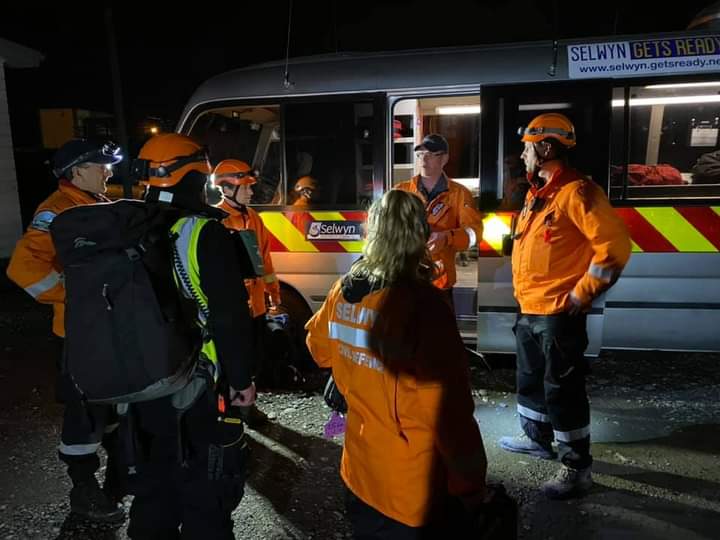 A group of people in orange high-viz vests and orange hard hat helmets stand in a circle in the dark talking next to a van with a yellow and red emergency stripe down the side