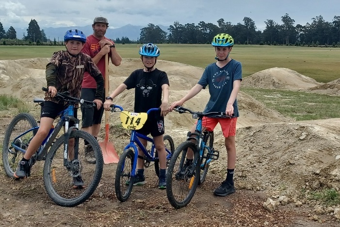 Three children on bicycles standing in front of a man in a red shirt leaning on a spade, all in front of a pump track