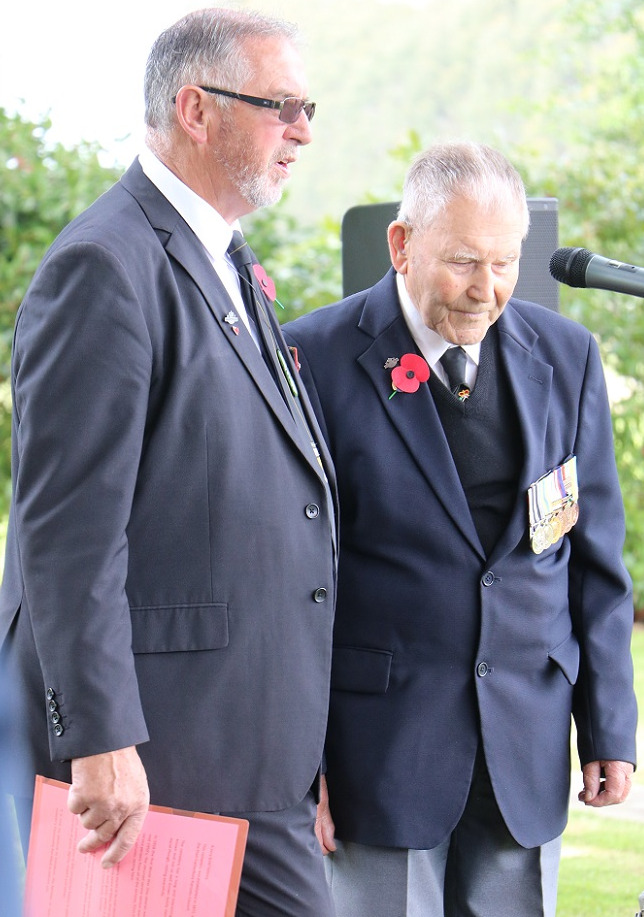 A grey haired man in a blue suit stands next to an elderly man in a suit wearing a poppy and a row of medals