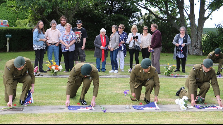 Four soldiers in dress uniforms kneel and lift flags from plaques while family members look at plaques on a row behind