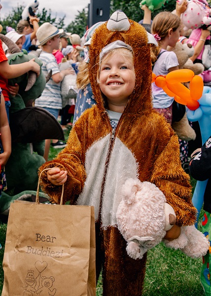 A little girl in a teddy bear costume carrying a teddy bear and a paper bag