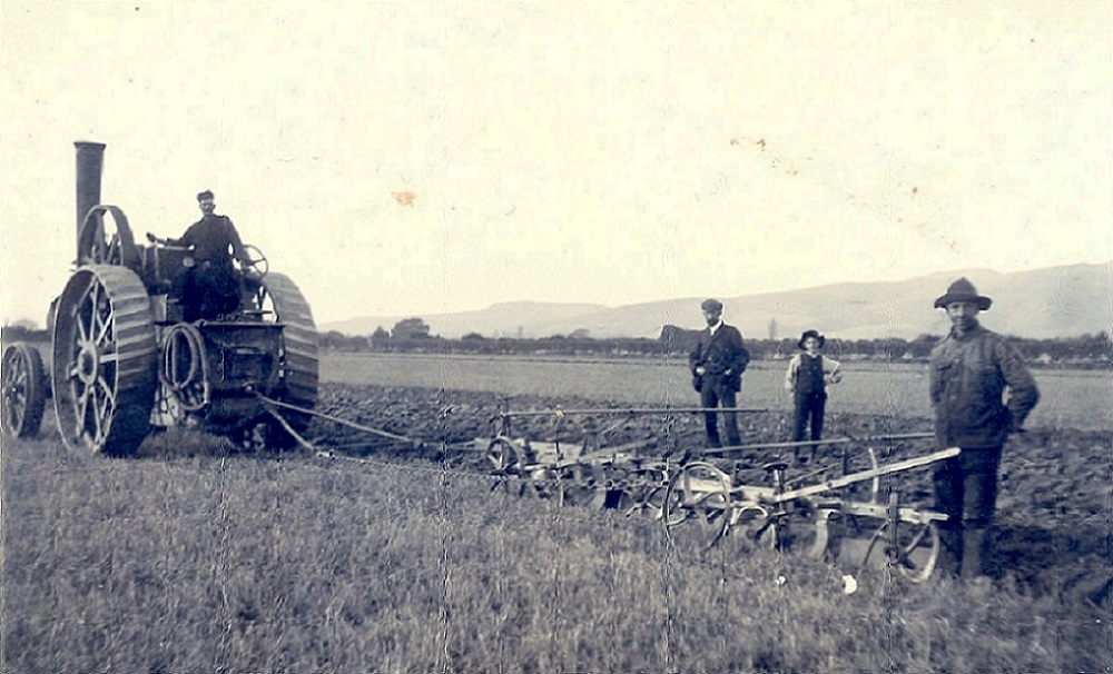 A historic black and white photo of a man on a tractor halfway through a ploughed field, while two men and a child stand in the ploughed area with their hands on their hips