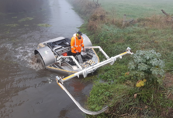 A boat in the river with its weed cutter on the front reaching up onto the bank