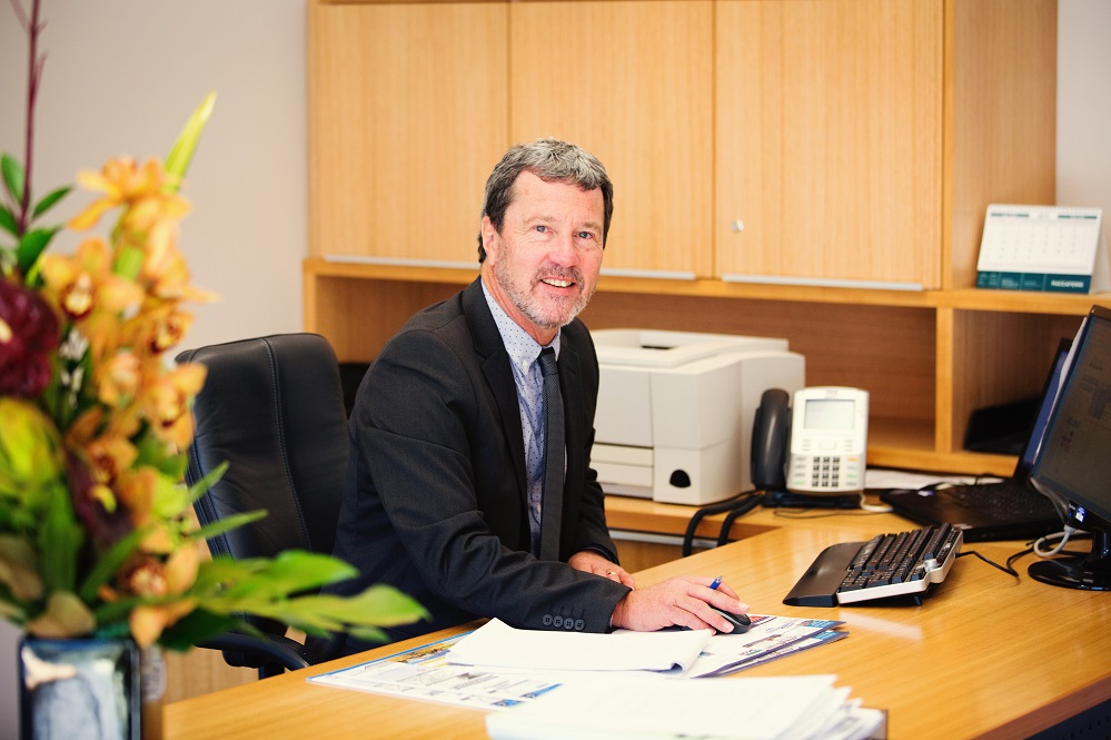 Young looking David Ward at desk with printer, cupboards, old phone behind him