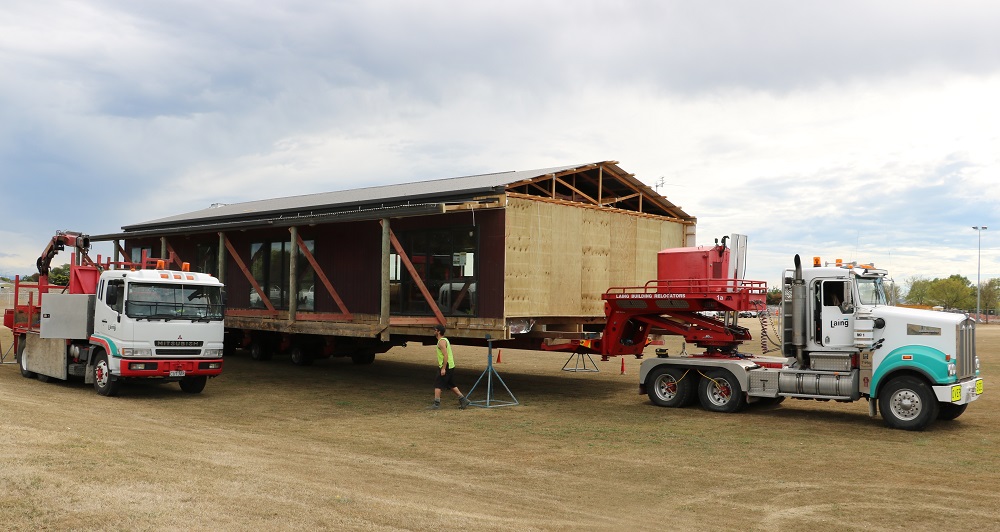 Half a single story brown building sits on the back of a green and white moving truck