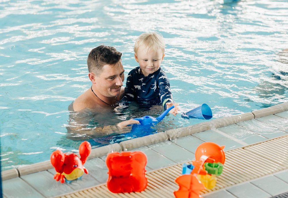 A dad and a young boy in the pool near the edge playing with a selection of toys including a blue watering can