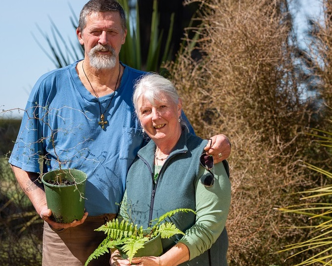 Stephen Clarke, in a blue shirt t-shirt stands with his arm around Lesley Barlow in a green gilet and shirt, while they both hold plants in pots