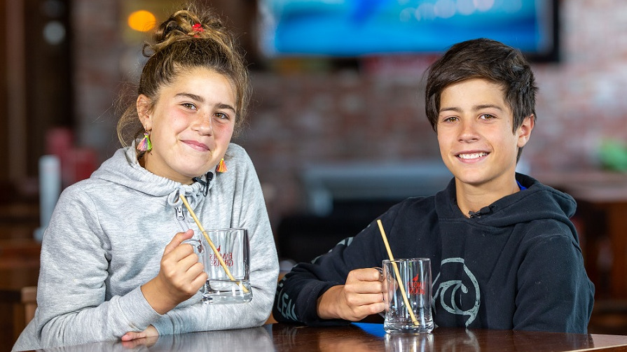 Girl with brown hair and grey hoodie and boy with black hair and black hoodie sit at a wooden table in a bar holding beer glasses with metal straws in them