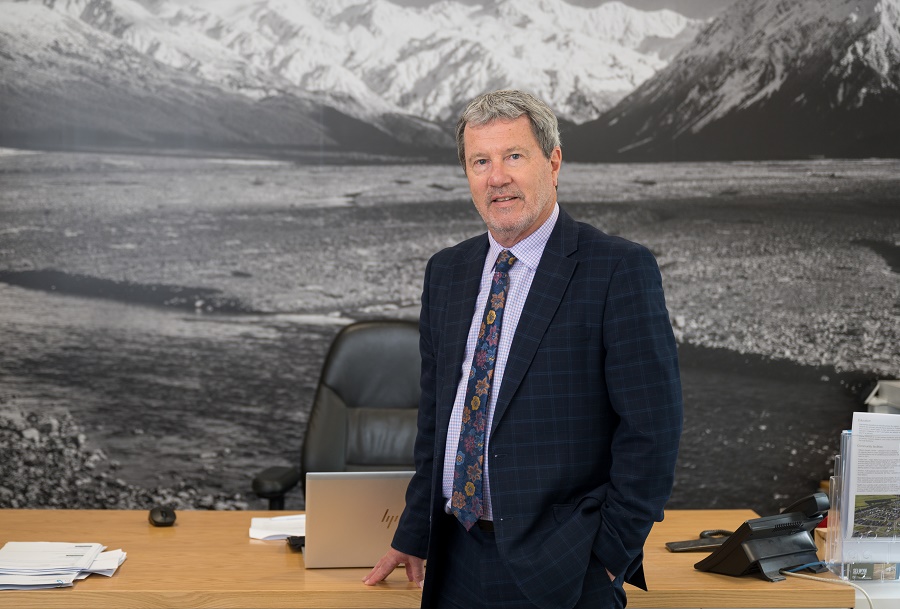 David Ward in a suit standing in an office with a black and white image of a high country river behind him