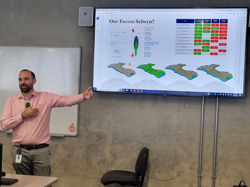 A balding man with a beard and wearing a pink shirt teaching in front of a screen with information about the Future Selwyn strategy