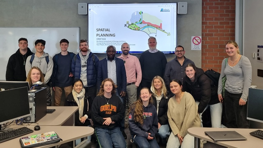 A group of students and staff lined up in two horizontal lines one behind the other, in front of a screen with an image and text saying spatial planning