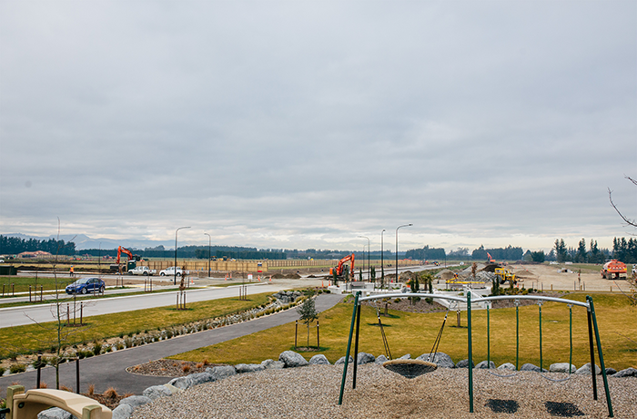 Residential development site, swing set and playground in front, red digger in back with hilly backdrop, roads and green land