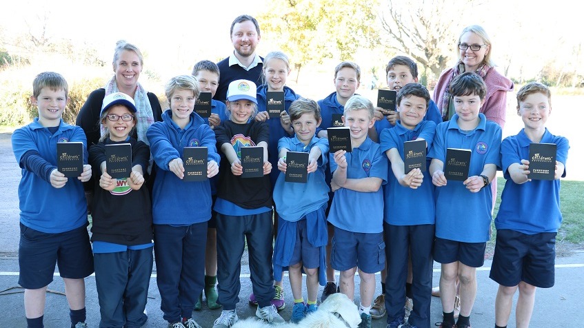 Children in two rows wearing blue school uniforms, two wearing white hats and black t-shirts with Selwyn District logo hold up small black books titled Children's University in front of two teachers and Mayor Sam Broughton
