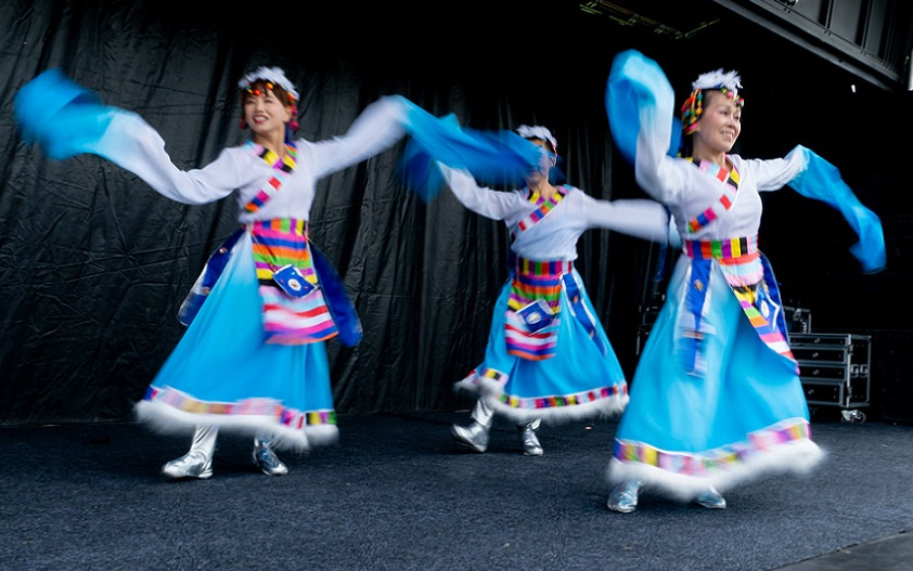 Three dancers in traditional outfits of blue with extra long sleeves, multi coloured checker trim and silver boots dance at CultureFest