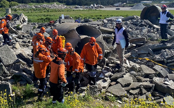 A group of people in orange flouro jackets and hard hats look through a pile of concrete rubble