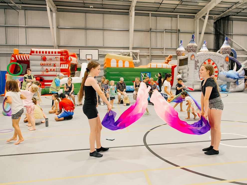 Two girls swish purple and pink scarves with other children behind playing with hoops, juggling clubs, scarves or on bouncy castles inside the Selwyn Sports Centre
