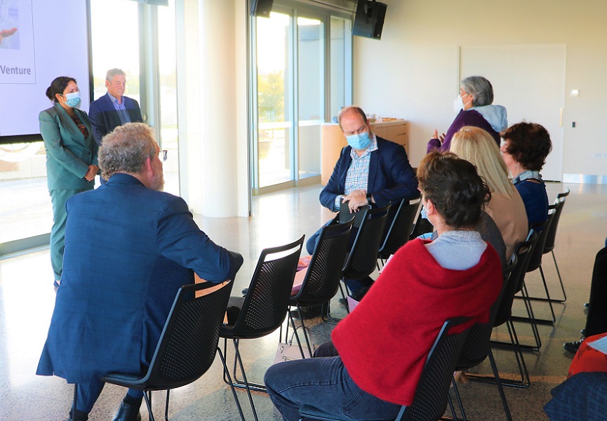 Minister Ayesha Verral and Dr Chris Kirk stand listening to a woman talk from a group of people seated in front of them
