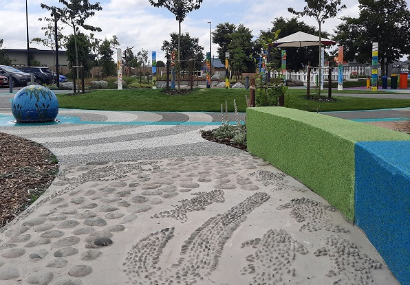 image of a pathway with raised fish and fishing motifs, a mosaic fountain feature, small grass hill and art poles in the sensory space