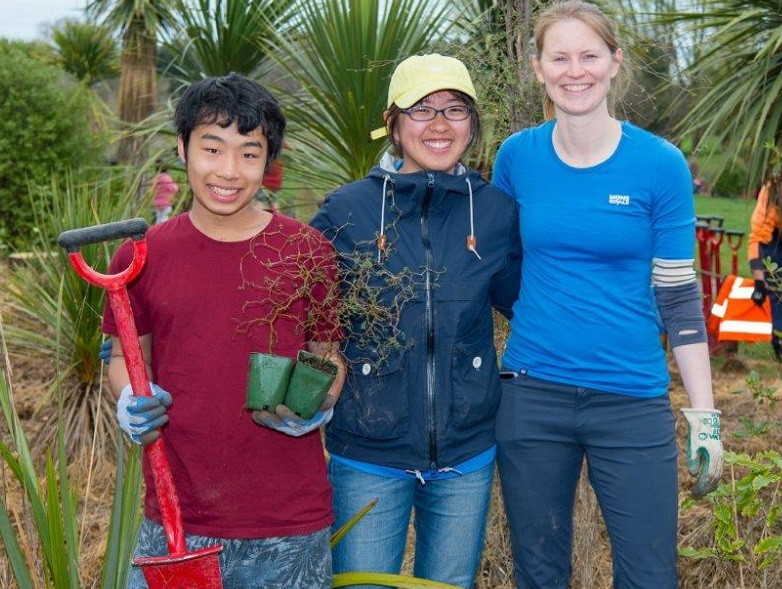 Two students and teacher, standing together, holding two seedlings and a spade