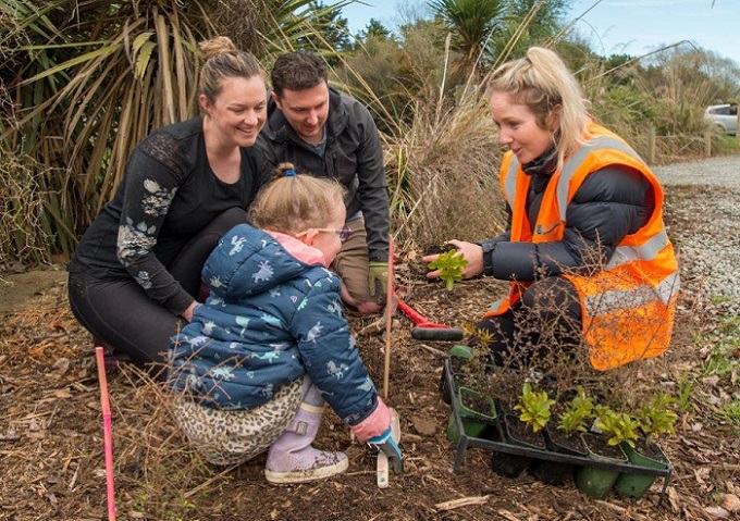 A mum, dad and young daughter talk to a woman in a high-viz jacket as they plant a tree together