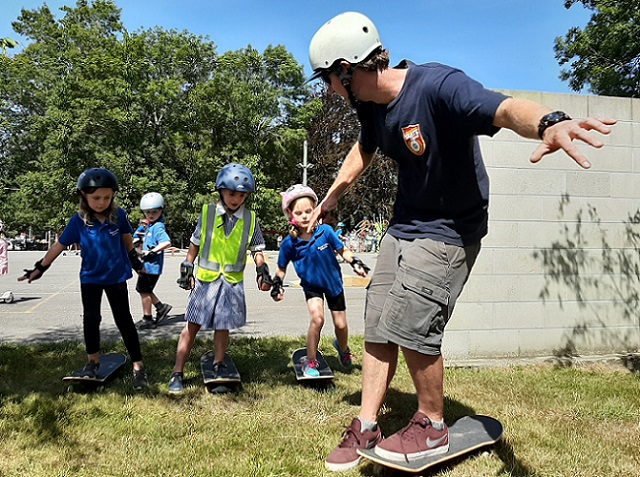 A man balances on a skateboard with his arms out wide, looking behind and talking to three primary school girls in uniform and helmets standing on skateboards