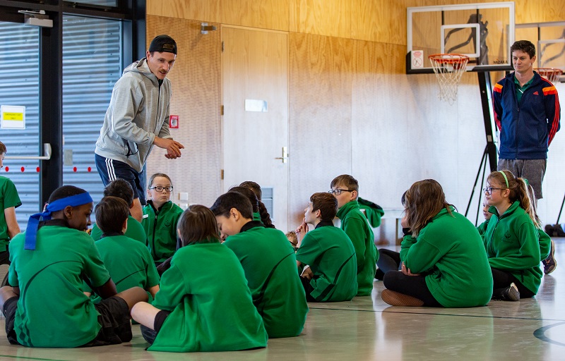 A man in a backwards black baseball cap, grey hoodie and blue sport shorts talks to a group of school children in green jerseys sitting on a gym floor, watched by a brown haired man in a blue tracksuit top with red striped sleeves