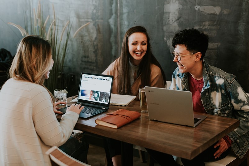 Three women laugh together while working on computers round a desk